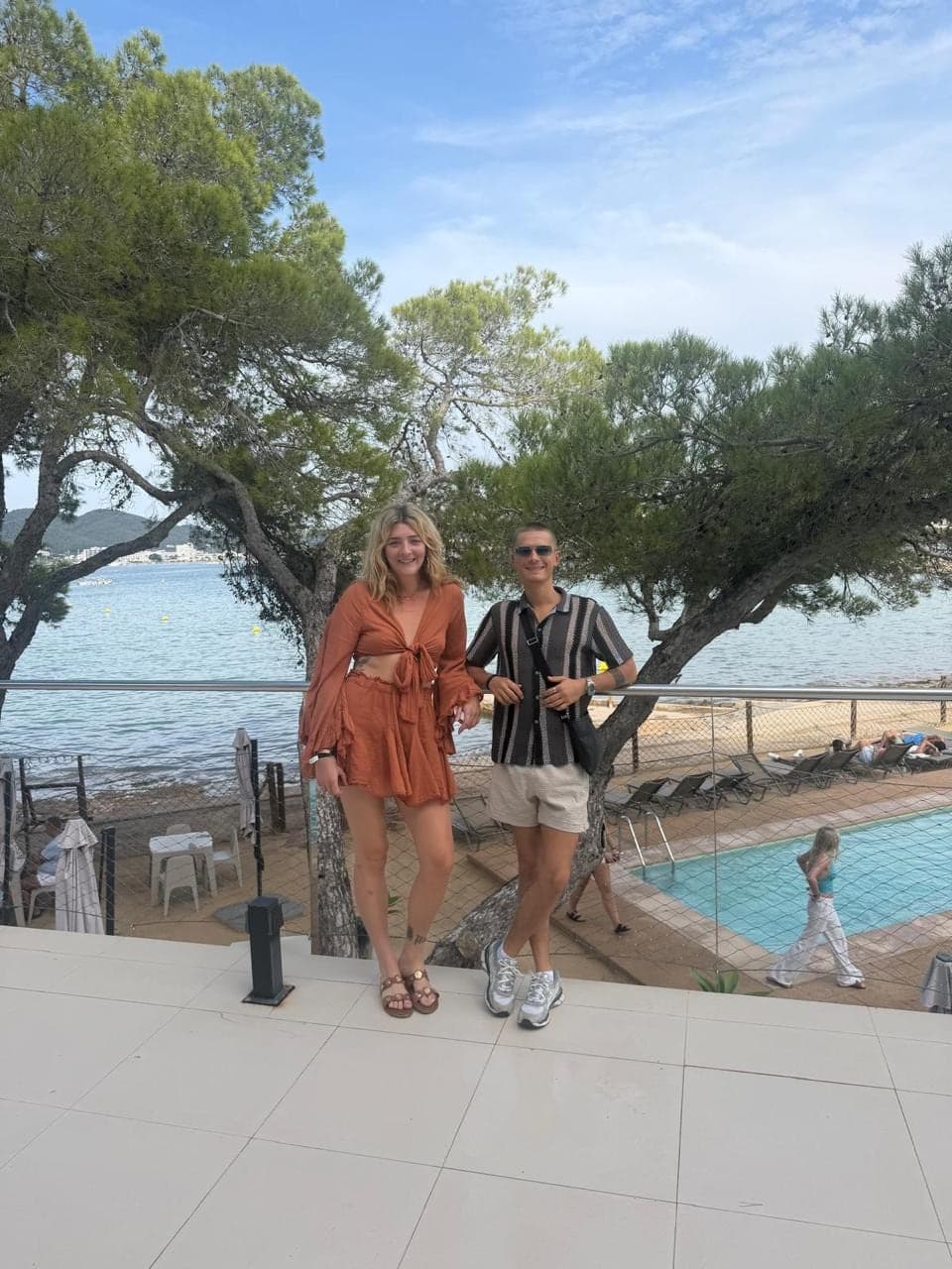 The two founders standing together on a sunlit terrace above the sea, pine trees behind them.